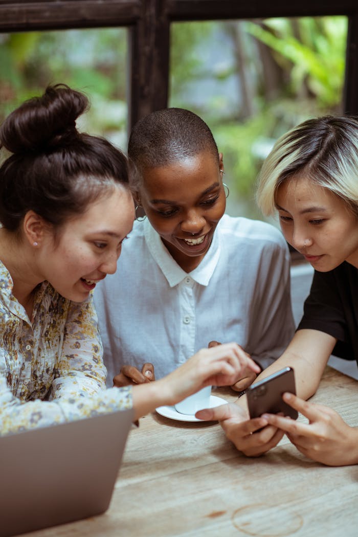 about-bg Three diverse women smile while using a smartphone in a cozy indoor setting.